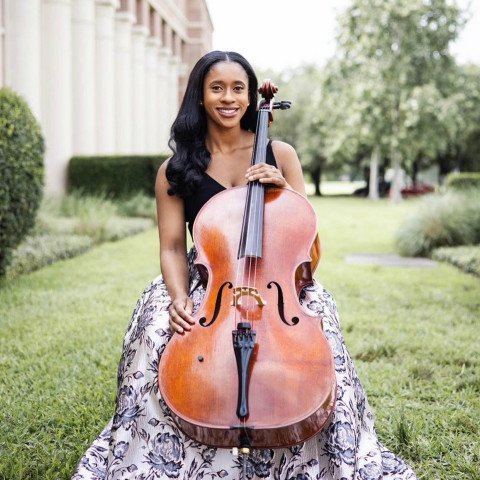 A Black woman wearing a floral dress, sitting in a park and holding a cello.