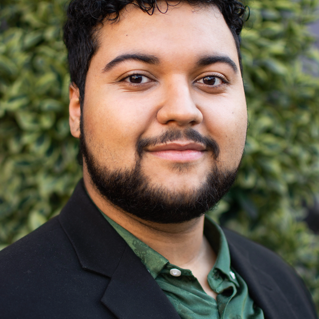 A headshot of a man with facial hair and mid-toned skin, wearing a green shirt and dark suit and smiling softly at the camera.