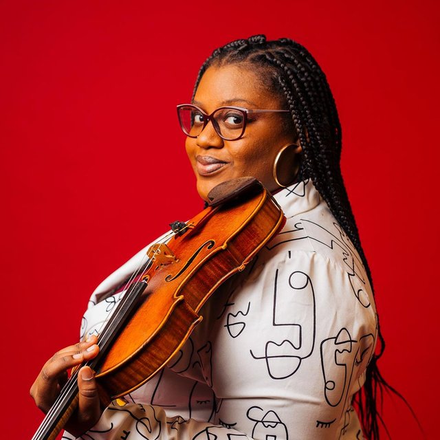 A black woman in profile with glasses and long braids stands in front of a bright red background holding a violin