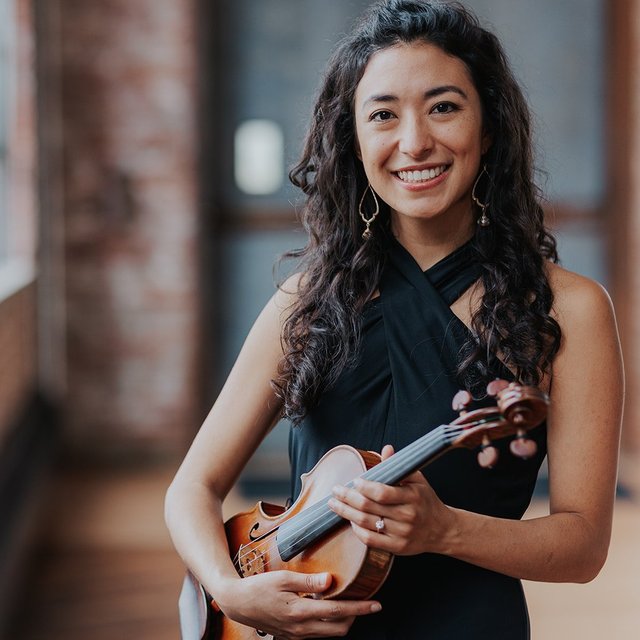 A woman with a bright smile and long dark hair looks directly into the camera, holding a violin.