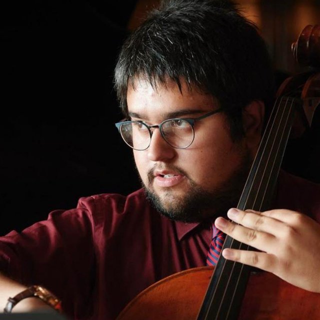 A close up on a man with dark hair wearing glasses and a red shirt playing the cello