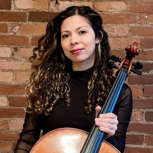A young woman with long dark curls stands with a cello and smiles subtly