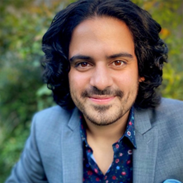 A Hispanic man with shoulder length dark hair and facial hair smiles at the camera