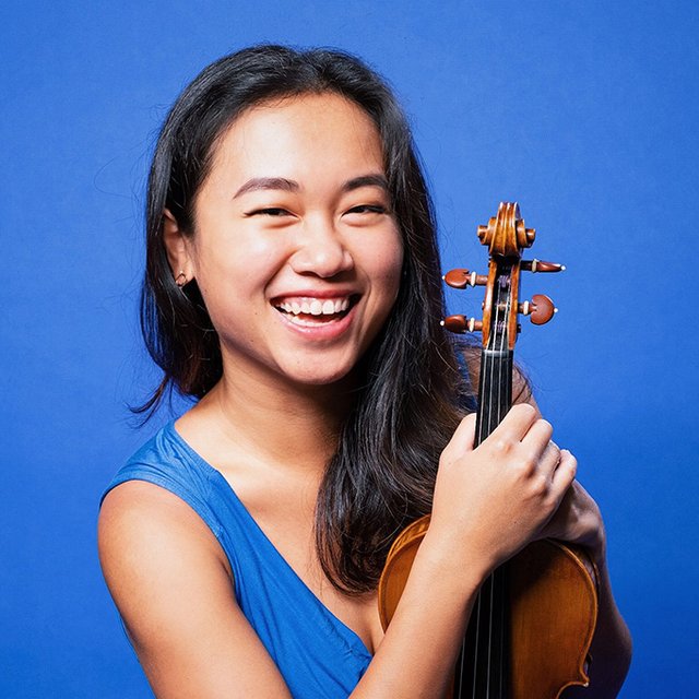 An Asian woman with long dark hair against a bright blue background holds a violin and smiles brightly