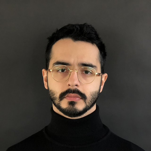 A young man with short dark hair & glasses looks at the camera with a serious expression
