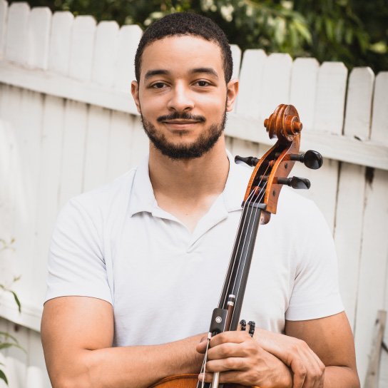 A man with facial hair and mid-toned skin, wearing a white shirt and holding a cello.