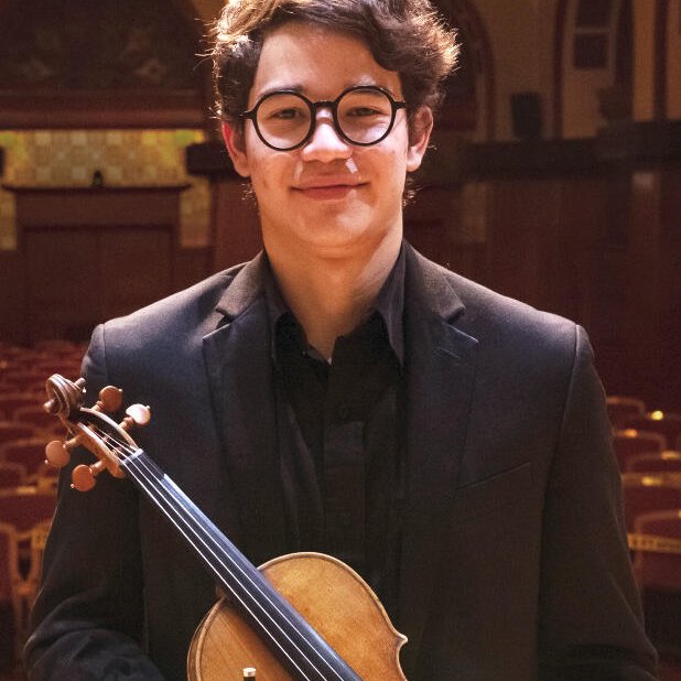a young man with round glasses and dark hair holds a violin