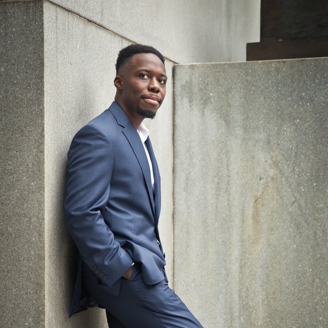 A full-body photo of a Black man in a blue suit, leaning against a concrete wall.
