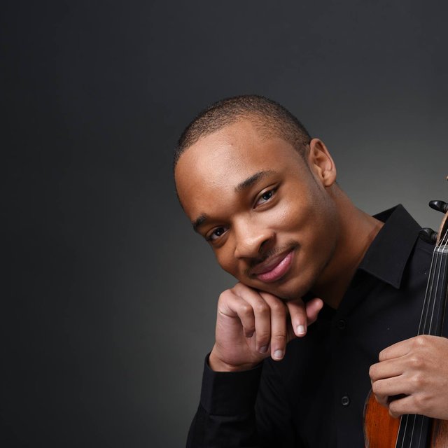 a young black man holding a violin looks at the camera with a subtle smile