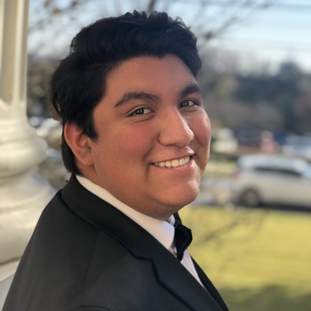A young man with medium tone skin and short dark hair wearing a suit looks over his shoulder and smiles