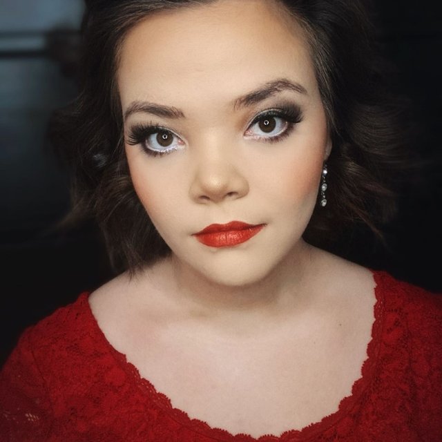 A close up headshot of an Asian woman wearing a red shirt and red lipstick looking directly into the camera