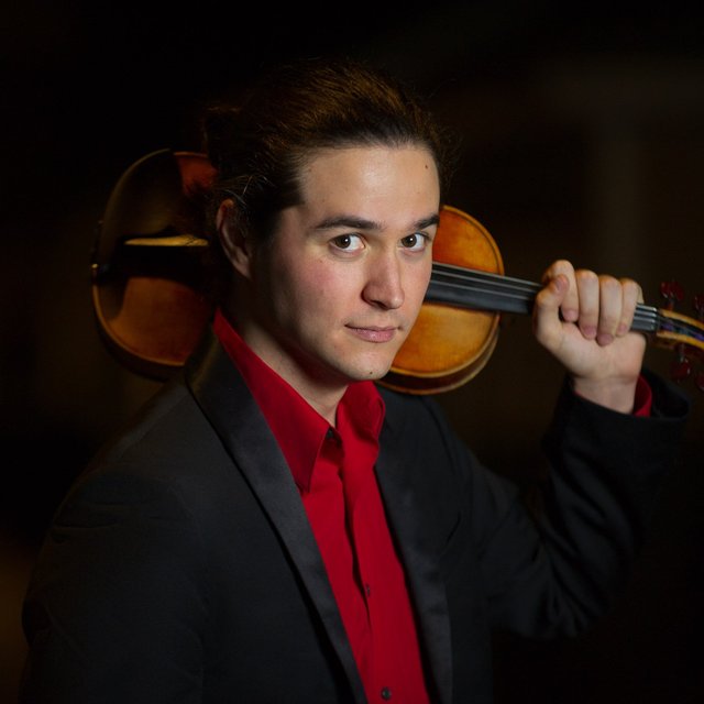 A man wearing a red shirt and black suit, balancing a violin over his shoulder.