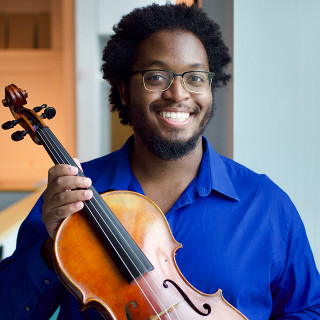 A young black man with glasses and a bright smile holds a viola and looks into the camera