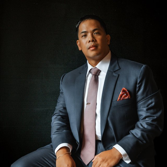 A Filipino man wearing a suit sits in front of a dark background and looks into the camera